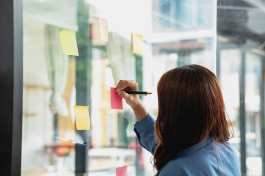 A female employee writes on a colorful notepad. Ethnic women working at startups brainstorm collaborative plans on glass wall stickers See less.