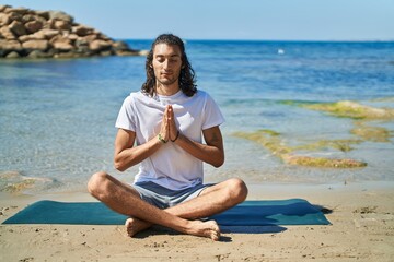 Fototapeta na wymiar Young hispanic man doing yoga exercise sitting on sand at beach