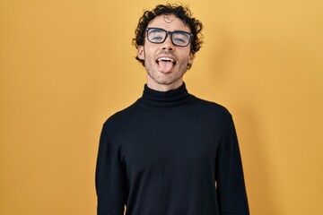 Hispanic man standing over yellow background sticking tongue out happy with funny expression. emotion concept.