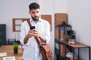 Hispanic man with beard using smartphone at the office in shock face, looking skeptical and...