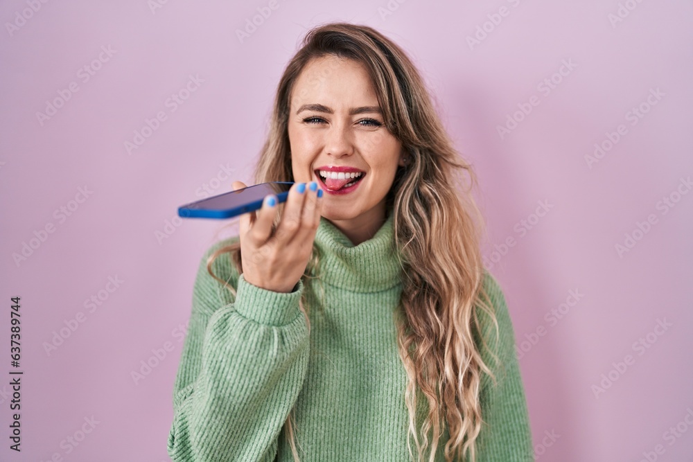 Poster young caucasian woman sending a voice message with smartphone sticking tongue out happy with funny e