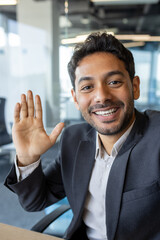 Vertical shot, young successful businessman looking into smartphone camera talking on video call using app on phone, smiling and having friendly conversation with friends and colleague inside office.