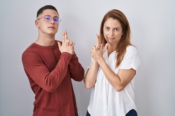 Mother and son standing together over isolated background holding symbolic gun with hand gesture, playing killing shooting weapons, angry face