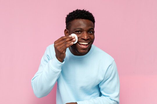 african american man in blue sweater uses cotton pads and smiles on pink isolated background