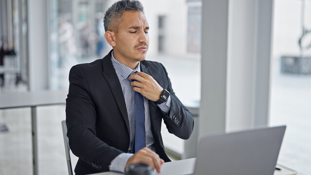 Young hispanic man business worker using laptop touching tie at office
