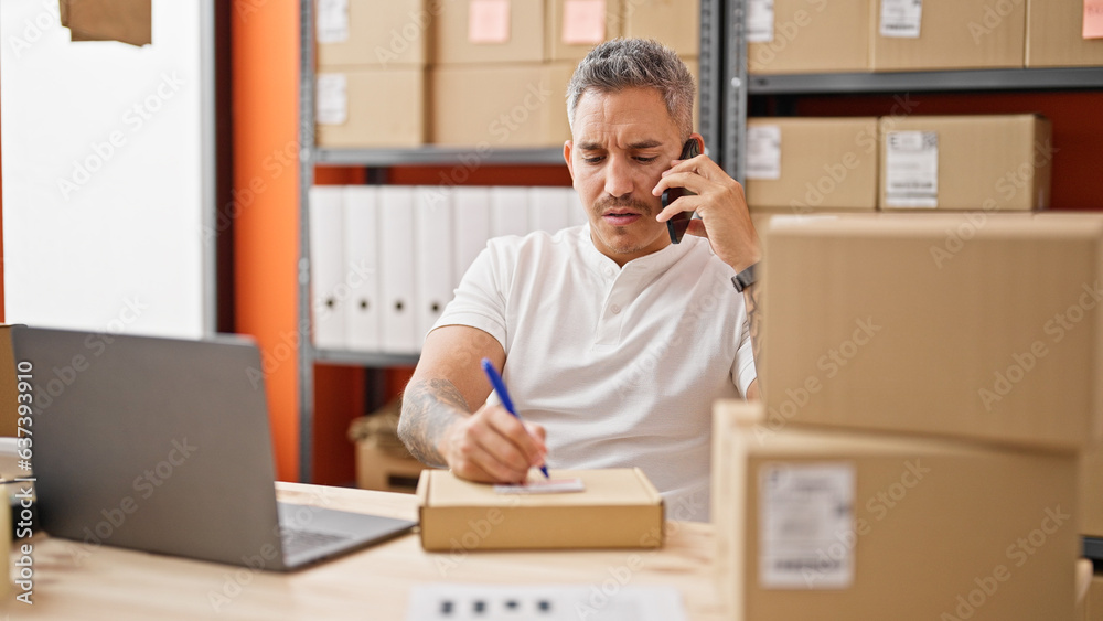 Sticker young hispanic man ecommerce business worker talking on smartphone writing on package at office