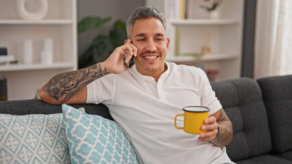 Young hispanic man talking on smartphone drinking coffee at home