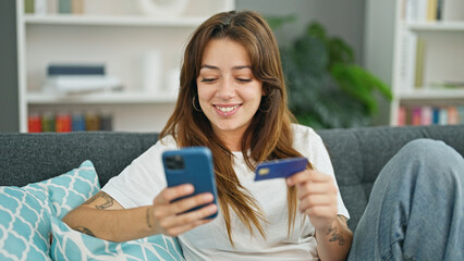 Young beautiful hispanic woman shopping with smartphone and credit card sitting on sofa at home