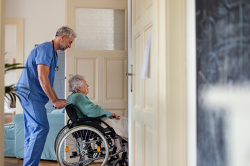 Caregiver pushing senior woman in a wheelchair, helping patient to move in her home.