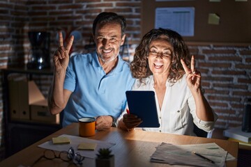 Middle age hispanic couple using touchpad sitting on the table at night smiling looking to the camera showing fingers doing victory sign. number two.