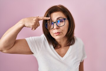 Middle age hispanic woman standing over pink background pointing unhappy to pimple on forehead, ugly infection of blackhead. acne and skin problem