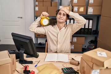 Young man working at small business ecommerce holding alarm clock stressed and frustrated with hand...