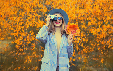 Autumn color style outfit, stylish young woman photographer with film camera and yellow maple leaves wearing round hat and coat in the park