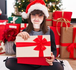 Adorable hispanic girl unpacking gift sitting by christmas tree at home