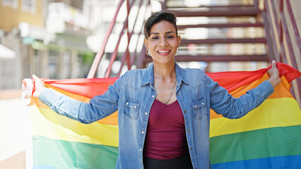 Young beautiful hispanic woman smiling confident holding rainbow flag at street
