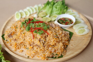 Pork fried rice with egg topped with coriander leaves and sliced ​​red chili. Fried rice served on a wooden plate decorated with sliced ​​cucumbers, lime wedges, spring onions and chili fish sauce.
