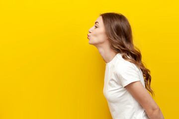 young cute girl sends air kiss to the side on yellow isolated background, woman flirts and shows...