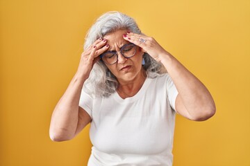 Middle age woman with grey hair standing over yellow background with hand on head for pain in head...