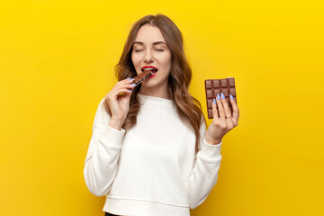 young girl eats and bites big chocolate bar and smiles and rejoices on yellow isolated background