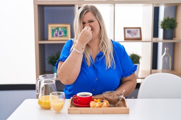Obraz na płótnie Canvas Caucasian plus size woman eating breakfast at home smelling something stinky and disgusting, intolerable smell, holding breath with fingers on nose. bad smell