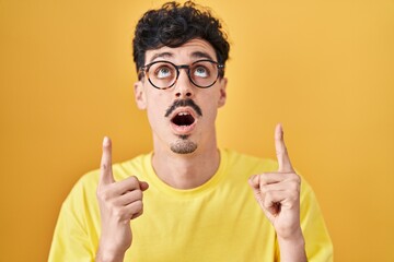 Hispanic man wearing glasses standing over yellow background amazed and surprised looking up and pointing with fingers and raised arms.