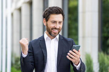 A young businessman man in a business suit stands outside on the street and looks at the phone,...