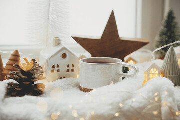 Winter hygge. Stylish cup of tea with modern cute christmas houses, pine cone, wooden star and tree, golden lights on soft warm blanket on windowsill. Christmas cozy still life. Merry Christmas!