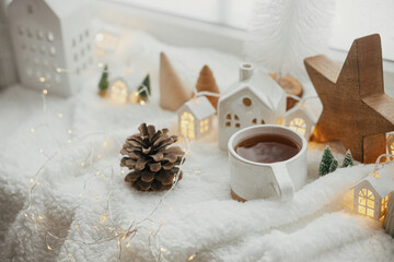 Obraz na płótnie Canvas Winter hygge. Stylish cup of tea with modern cute christmas houses, pine cone, wooden star and tree, golden lights on soft warm blanket on windowsill. Christmas cozy still life. Merry Christmas!