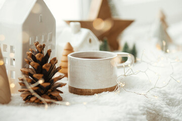 Obraz na płótnie Canvas Cozy winter still life. Stylish cup of tea with modern christmas decoration, pine cone, wooden star and tree, golden lights on soft warm blanket on windowsill. Merry Christmas and Happy holidays