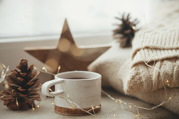 Cozy winter still life. Stylish cup of tea with cozy knitted sweater, pine cone, wooden star and...