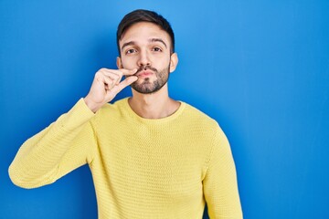 Hispanic man standing over blue background mouth and lips shut as zip with fingers. secret and...