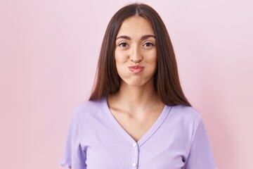Young hispanic woman with long hair standing over pink background puffing cheeks with funny face. mouth inflated with air, crazy expression. - obrazy, fototapety, plakaty