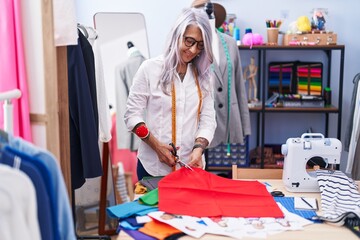 Middle age grey-haired woman tailor smiling confident cutting cloth at tailor shop