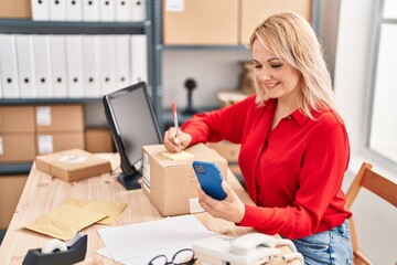 Young blonde woman ecommerce business worker writing on package using smartphone at office