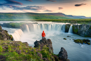 Godafoss waterfall flowing with colorful sunset sky and male tourist standing on cliff in summer at...