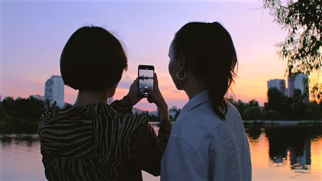 Women take photos of lake with mobile phone. Beautiful girls taking video on mobile phone of a sunset, female couple, friends in summer evening
