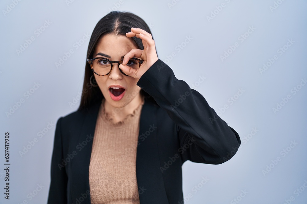 Poster young brunette woman standing over blue background doing ok gesture shocked with surprised face, eye