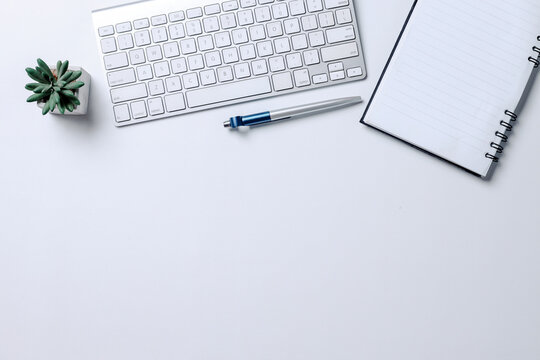 Top view of keyboard computer, notebook, pen and succulent on white desk with blank space for text or ads
