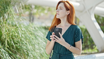 Young redhead woman using smartphone with serious expression at park