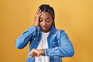 African american woman with braids standing over yellow background looking at the watch time...