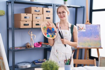 Young caucasian woman artist smiling confident holding paintbrushes and palette at art studio