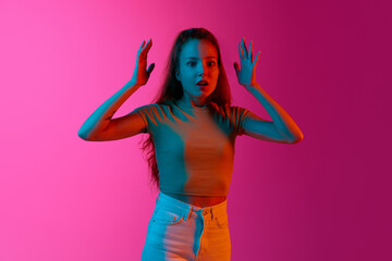 Young emotive girl raising hands up, standing with widely open eyes, shocked face against pink studio background in neon light. Concept of human emotions, fashion, beauty, lifestyle, youth, ad