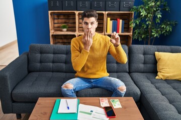 Young hispanic man holding bitcoin virtual money covering mouth with hand, shocked and afraid for...
