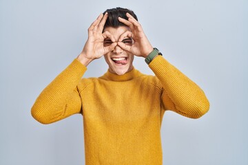 Young hispanic man standing over blue background doing ok gesture like binoculars sticking tongue out, eyes looking through fingers. crazy expression.