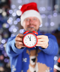 Bearded smiling man wearing warm blue sweater with traditional deer hold arm red alarm clock within...