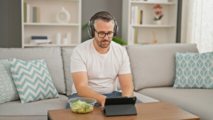 Grey-haired man eating salad using touchpad and headphones at home