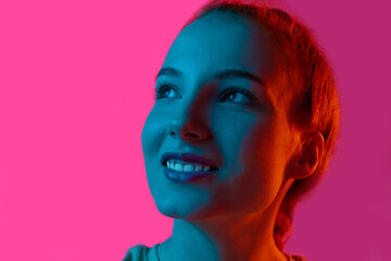 Close-up portrait of young smiling girl looking upwards against pink studio background in neon light. Concept of human emotions, fashion, beauty, lifestyle, youth, ad