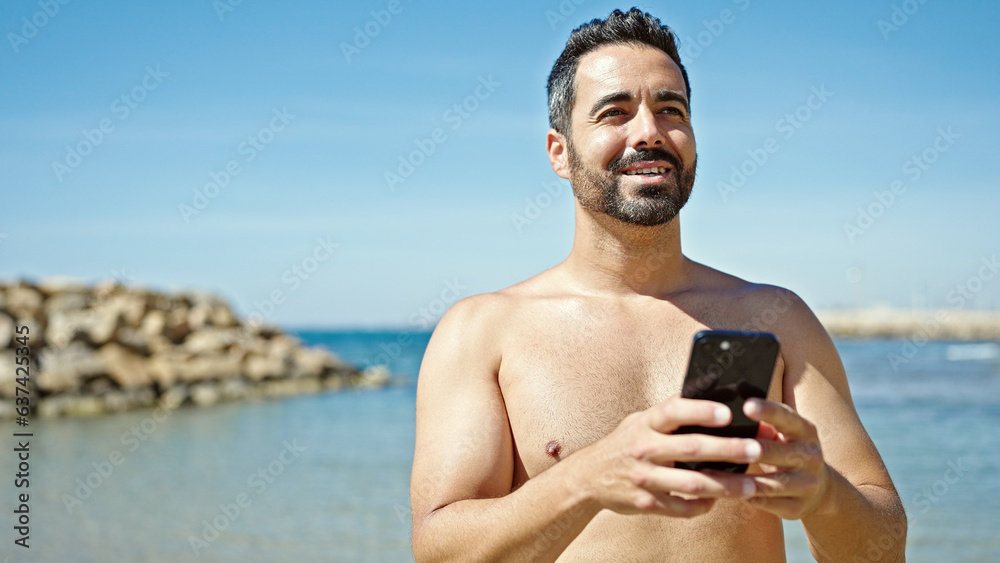 Sticker Young hispanic man tourist using smartphone standing shirtless at the beach