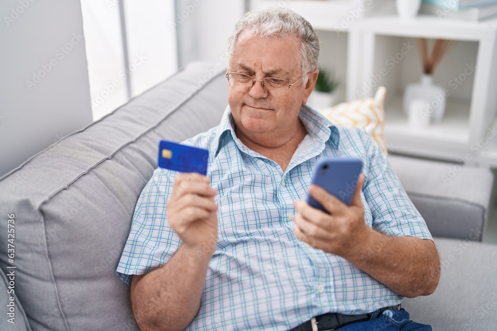 Wall mural Middle age grey-haired man using smartphone and credit card sitting on sofa at home