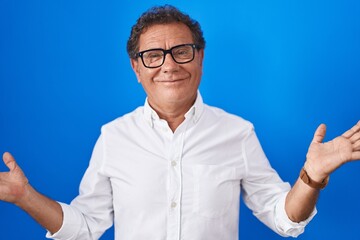 Middle age hispanic man standing over blue background clueless and confused expression with arms...